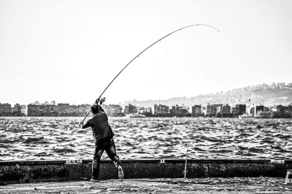 A man casting a fishing line by the sea with a cityscape in the background.
